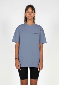 Kortærmet, oversized t-shirt i blødt blåt bomuld med et lille sort grafisk design på venstre bryst. Bæres med sorte cykelshorts.