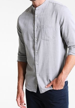 Mężczyzna ubrany w jasno szarą koszulę z kołnierzem mandaryńskim zapinaną na guziki, z kieszenią na piersi oraz ciemnoniebieskie spodnie, z jedną ręką w kieszeni.