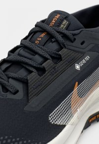 Gros plan sur une chaussure de course Nike Pegasus Trail 5 Gore-Tex noire avec des lacets texturés, des étiquettes de marque et un swoosh orange sur une semelle blanche.