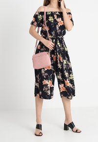 Tuta floreale nera con design off-the-shoulder, vita stretta e gambe corte. Abbinato a una borsa a tracolla rosa e sandali neri con tacco a blocco.