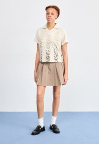 Kortärmad gräddfärgad polo med leopardmönster, matchad med en beige plisserad kjol och svarta skor, bärs med vita strumpor.