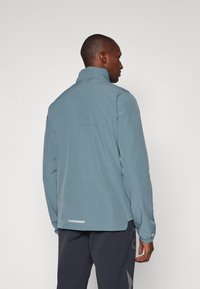 Veste pour homme en tissu bleu clair, avec un col montant, des manches longues et un design au dos avec une couture horizontale et des détails réfléchissants.