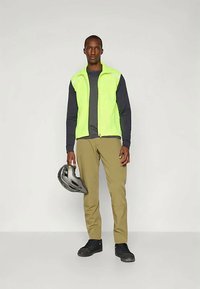 Atstarojošs neona zaļš veste, tumši zila garenā piedurkne krekls, khaki bikses, melnas kurpes, turot pelēku ķiveri, stāvot uz neitrāla fona.