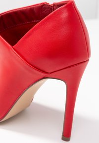 Zapato de tacón alto de cuero rojo con punta en forma de punta y tacón de aguja delgado. Textura suave y detalle de costuras mínimas en la parte superior.