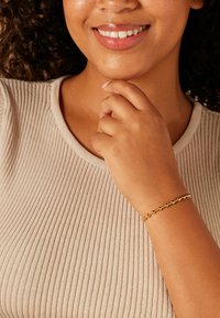 Gold beaded bracelet with spherical accents, worn on a wrist against a textured beige ribbed top. Subtle shine on the beads.