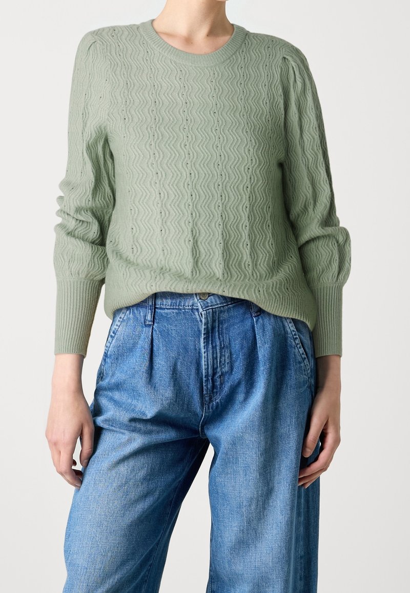 Personne portant un pull en tricot texturé vert clair avec un motif en zigzag et un jean ample taille haute bleu clair, debout devant un fond uni.