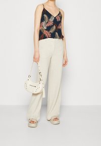 Nainen, jolla on päällään beige leveälahkeiset housut, tummansininen toppi, jossa on vaaleanpunaisia ja beigejä lehtikuvioita, beige platform-sandaalit, ja hän pitää kädessään beigeä käsilaukkua.