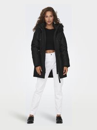 Black parka with a fur-lined hood, featuring a zip closure and side pockets. Worn over a black crop top and white pants, with black shoes.