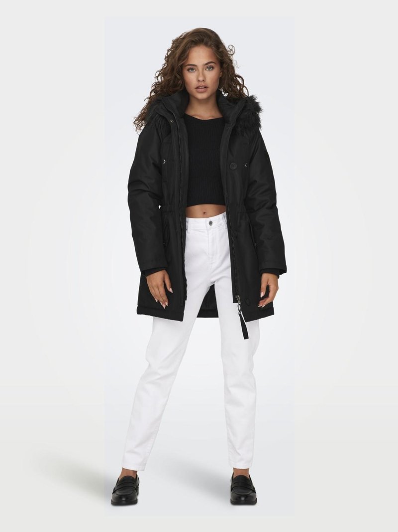 Parka noire avec une capuche en fourrure, dotée d'une fermeture éclair et de poches latérales. Portée sur un top court noir et un pantalon blanc, avec des chaussures noires.