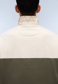 Genser med krage, med en kremfarget overdel, olivengrønn underdel, og oransje diamantsting på kragen. Glatt tekstur.