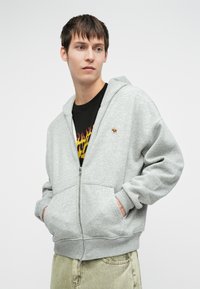 Siva zip-up kapuca iz flisa, s kenguru žepom, rebrastim manžetama i izveznim logotipom na prsima, nosi se preko crne majice.