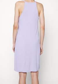 Persoon in een mouwloze, lichtpaarse jurk tot de knie, gezien van achteren, met een kleine tattoo op hun linkerarm.