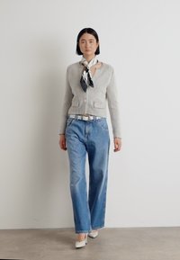 Grijze textuur cardigan met twee zakken, gedragen over lichtblauwe wijde spijkerbroeken. Een zwart-wit sjaal gestrikt om de nek; model in witte hakken.