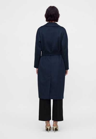 Personne aux cheveux courts et foncés portant un manteau bleu marine ceinturé, un pantalon noir à jambes larges et des chaussures à talons beiges, vue de dos sur un fond blanc.