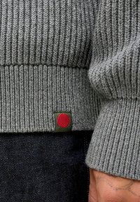 Tuvumā redzam pelēku ribotu džemperu piedurkni ar mazu zaļu birku, uz kuras ir sarkans aplis, kas valkāts kopā ar tumšu džinsu audumu un redzamu roku tetovējumu.