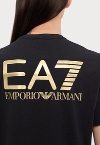 Juodas trumpas rankovėmis marškinėliai su ryškiomis auksinėmis raidėmis "EA7 EMPORIO ARMANI" nugarėlėje, su unikaliu dizainu ir tekstūra.