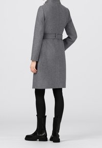 Grå ullkappa med hög krage och bälte bak, skräddarsydd passform, långa ärmar, parat med svarta leggings och kraftiga svarta stövlar.