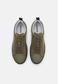 Ελαιόχρωμα αθλητικά παπούτσια με υφή στην επιφάνεια, στρογγυλή μύτη, καουτσούκ σόλα και μαύρες λεπτομέρειες. Συμπεριλαμβάνονται μονοχρωμαί κορδόνια και ανάγλυφο λογότυπο στη γλώσσα.