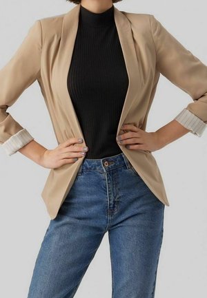 Osoba ubrana w beżową marynarkę na czarną prążkowaną bluzkę, z rękami na biodrach, w połączeniu z wysokim stanem niebieskich dżinsów na jednolitym tle.