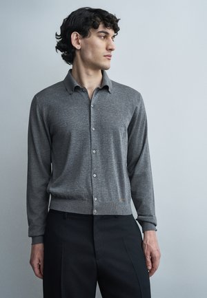 Νεαρός άνδρας με σγουρά σκουρόχρωμα μαλλιά, φορώντας εφαρμοστό γκρι πουλόβερ με κουμπιά και μαύρο παντελόνι, κοιτάζοντας προς το πλάι σε απλό φόντο.