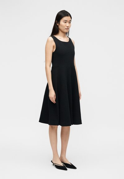 Γυναίκα που στέκεται με μαύρο αμάνικο φόρεμα μέχρι το γόνατο και μαύρες μυτερές γόβες με λουρί, μπροστά σε ανοιχτόχρωμο, απλό φόντο.