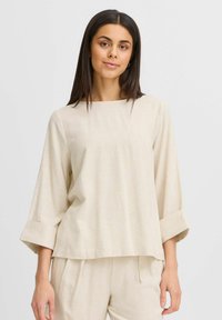 Haut beige, ample, en tissu texturé. Présente un décolleté carré et des manches trois-quarts avec des poignets repliés.