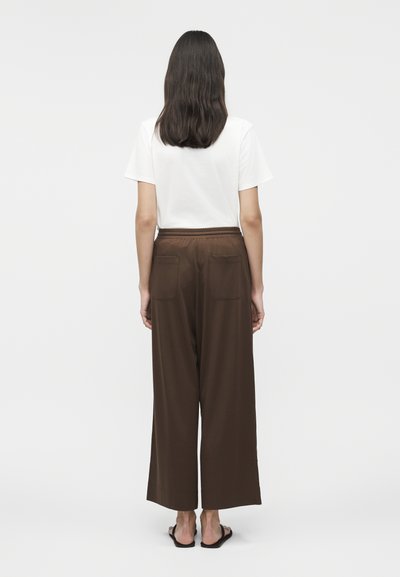 Γυναίκα με σκούρα μαλλιά μέχρι τον ώμο, γυρισμένη πλάτη, φοράει λευκή κοντομάνικη μπλούζα, καφέ παντελόνι με φαρδιά μπατζάκια και πίσω τσέπες, και μαύρα σανδάλια.