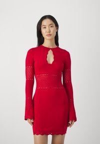 Donna che indossa un abito rosso aderente con maniche lunghe, aperture a forma di diamante sulle maniche e in vita, e scollatura a chiave.