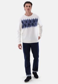 Weißer Pullover mit blauem geometrischem Muster, kombiniert mit dunkelblauen Jeans. Der Pullover hat einen runden Ausschnitt und gerippte Bündchen.