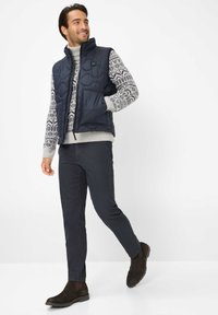 Marineblauer Steppweste über einem grauen gemusterten Pullover, dunklen Hosen und braunen Stiefeletten. Die Weste verfügt über einen Reißverschluss und eine Fronttasche.