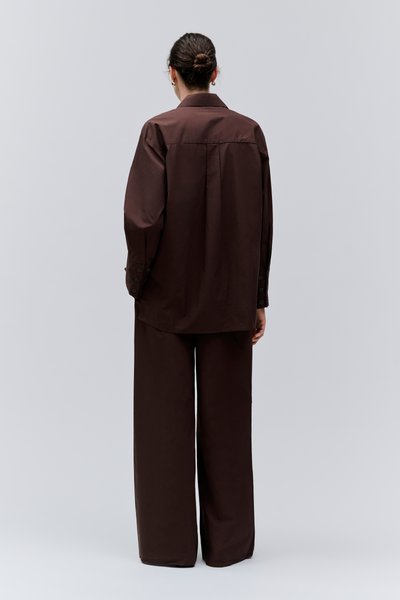 Personne avec les cheveux en chignon, portant une chemise ample marron foncé à manches longues et un pantalon large, debout de dos sur un fond uni.