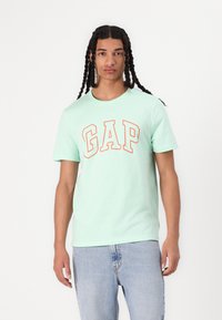 Camiseta de algodón verde menta con letras prominentes en naranja "GAP", mangas cortas y diseño de cuello redondo. Combinada con unos jeans de mezclilla azul claro.