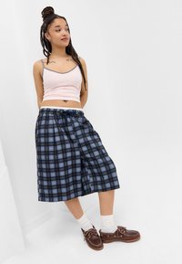 Blaue und schwarze Karottenhosen mit einem Kordelzugbund, kombiniert mit einem hellrosa Tanktop und braunen Bootsschuhen. Weiße Socken sind sichtbar.