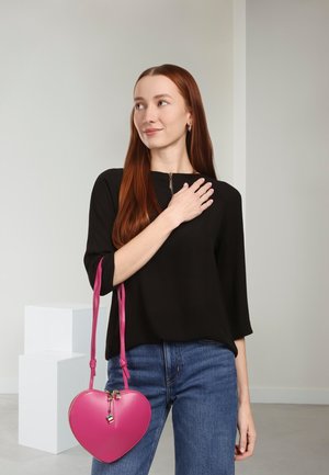 Sac à main en cuir rose en forme de cœur avec des bretelles ajustables, associé à un haut noir à manches longues et un jean bleu, sur un fond neutre.