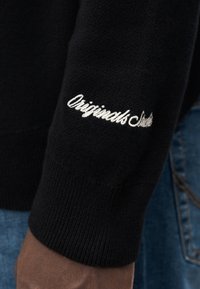 Μαύρο πλεκτό πουλόβερ με ριμπ μανσέτα και λευκό κεντημένο κείμενο που γράφει «Originals Studio». Με ομαλή υφή και εφαρμοστή σχεδίαση.