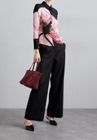 Donna che indossa pantaloni neri a gamba larga e una blusa nera con una grande stampa floreale rosa, tiene in mano una borsa bordeaux e indossa mule nere con tacco.