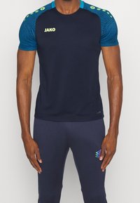 Marineblaues Sportshirt mit strukturiertem blauen Muster an den Schultern, grünem Logo und kurzen Ärmeln, kombiniert mit marineblauen Hosen mit buntem Akzent.