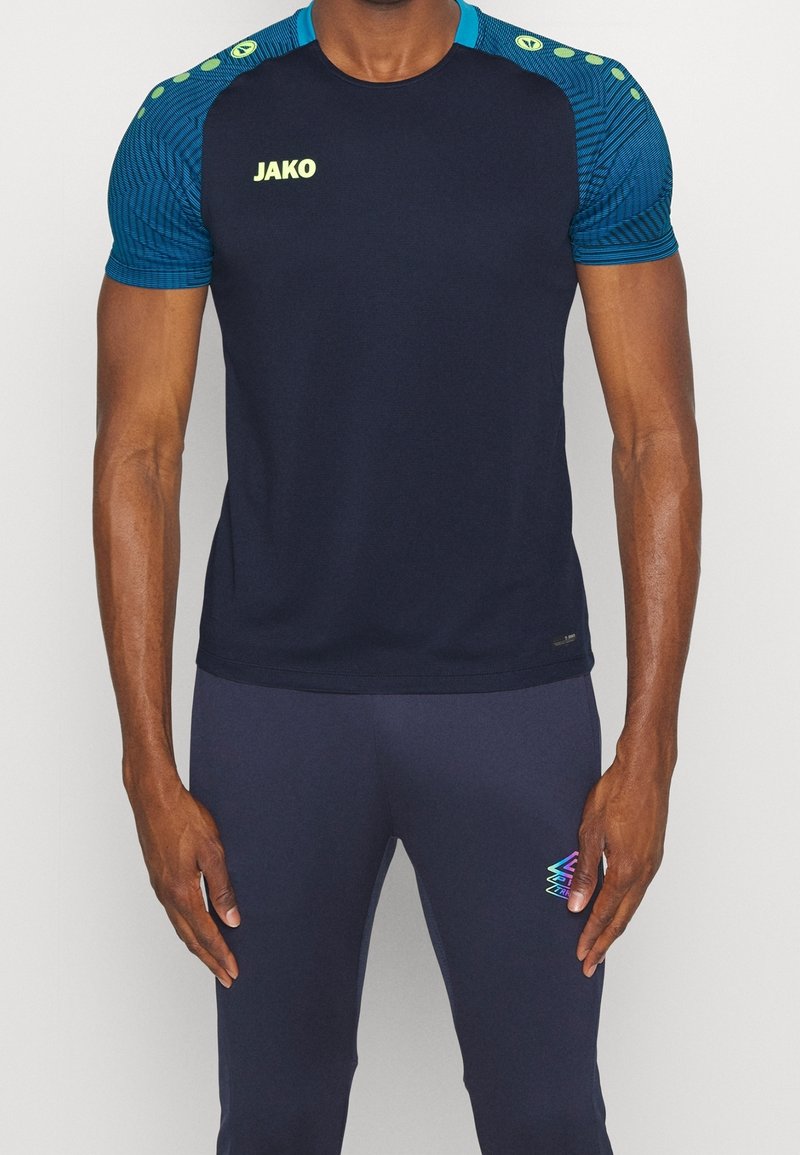 Marineblaues Sportshirt mit strukturiertem blauen Muster an den Schultern, grünem Logo und kurzen Ärmeln, kombiniert mit marineblauen Hosen mit buntem Akzent.