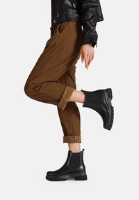 Stivali Chelsea neri impermeabili con accenti laterali in elastico testurizzato e suola in gomma spessa, abbinati a pantaloni in corduroy marroni.