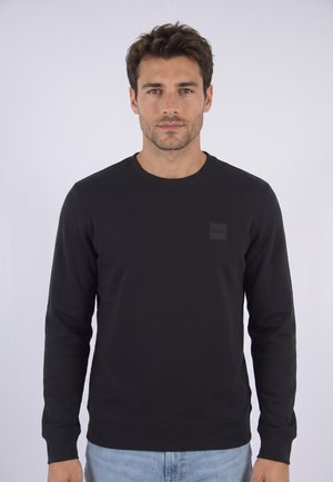 Homme aux cheveux bruns courts portant un t-shirt noir uni à manches longues et un jean bleu clair sur un fond clair uni.