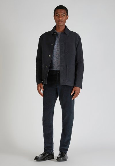 Mężczyzna ubrany w czarną kurtkę, wielokolorowy sweter, czarne spodnie sztruksowe i czarne skórzane buty, stojący na tle jasnoszarym.