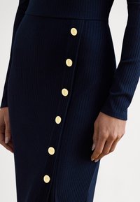 Marineblå ribbestrikket kjole med tettsittende design, lange ermer og gullknapper langs siden med inngravert dekor.