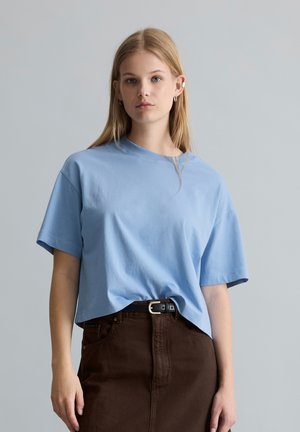 Junge Frau mit langen blonden Haaren, die ein hellblaues kurzärmeliges Oberteil und dunkelbraune Hose mit schwarzem Gürtel trägt, steht vor einem grauen Hintergrund.