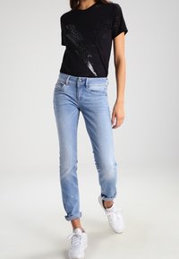 Černé tričko s opotřebovanými detaily a metalickým potiskem, v kombinaci se světle modrými slim-fit džíny a bílými teniskami. Jednoduché pozadí.