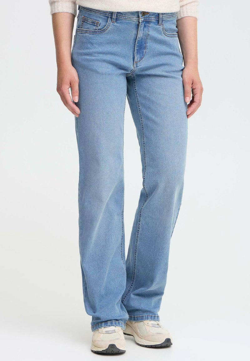 Jeans de denim azul claro con un diseño de pierna recta, cinco bolsillos y detalles de costura en contraste. Llevados con zapatillas beige.