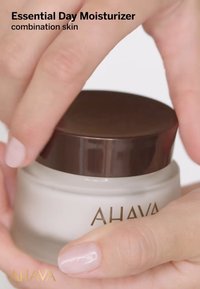 Tarro blanco con tapa marrón etiquetado como "AHAVA Essential Day Moisturizer". Las manos de una persona lo están abriendo, mostrando una textura suave y cremosa en su interior.