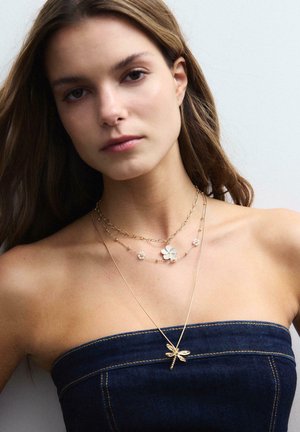 Mlada ženska, oblečena v top iz traperja brez naramnic in slojevne zlate ogrlice z obeskom v obliki cvetice in kačjega pastirja na enobarvnem ozadju.