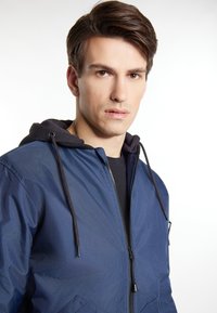 Giacca zip-up blu navy con cappuccio nero, caratterizzata da una texture liscia e tasche frontali. Il design include lacci e una silhouette aderente.