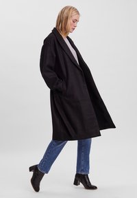 Cappotto di lana nera con vestibilità ampia, ampi revers e tasche laterali. Indossato sopra un maglione grigio, abbinato a jeans blu e stivali neri con tacco.
