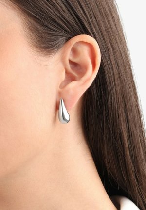 Pendiente en forma de lágrima plateado con una superficie lisa y reflectante, colgado de la lóbulos de la oreja, enmarcado por un cabello castaño liso.
